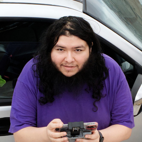 A man with long black hair and a beard, wearing a loose purple T-shirt, holds a drone controller and looks up toward the camera, standing beside a silver pickup truck.