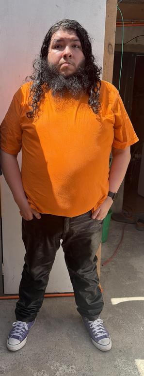 A person with long dark curly hair and a gray-streaked beard stands in a utility room, wearing a bright orange t-shirt, black leather pants, and purple sneakers.
---
Una persona con cabello largo, oscuro y rizado y una barba con mechones grises está de pie en un cuarto de servicio, vistiendo una polera naranja brillante, pantalones de cuero negros y zapatillas moradas.